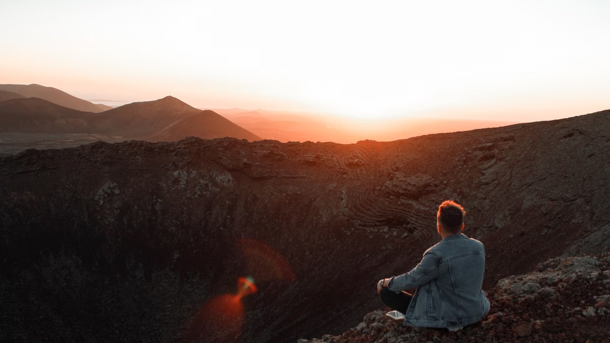 A person sits on a volcanic crater edge, meditating at sunset in La Oliva, Spain.