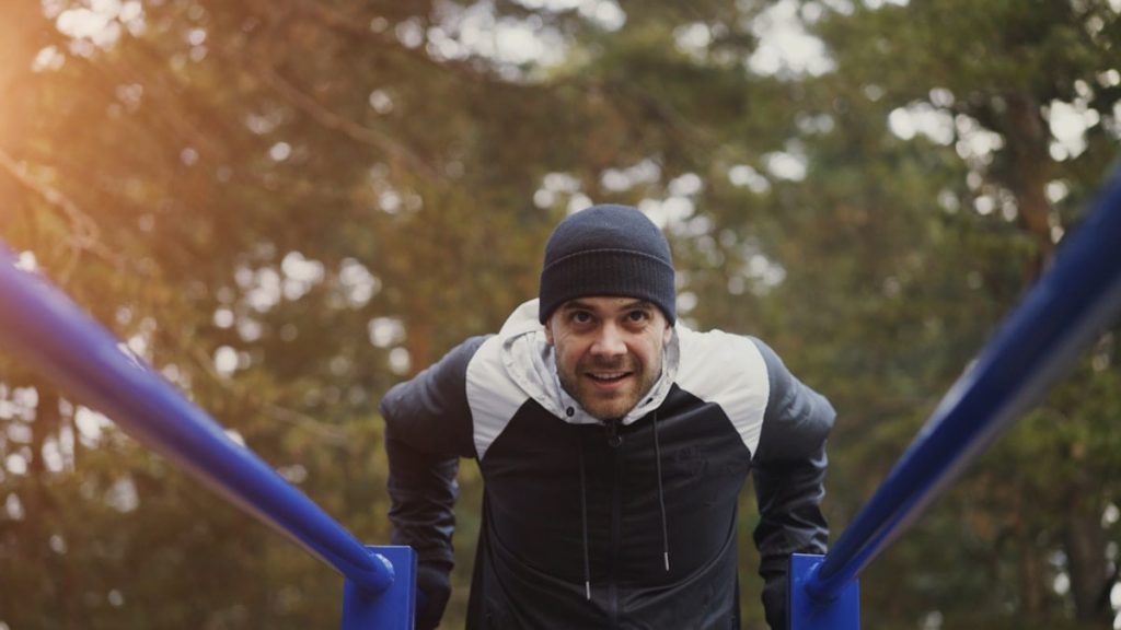 Young handsome athlete man doing push-ups exercise on bars in winter park outdoors